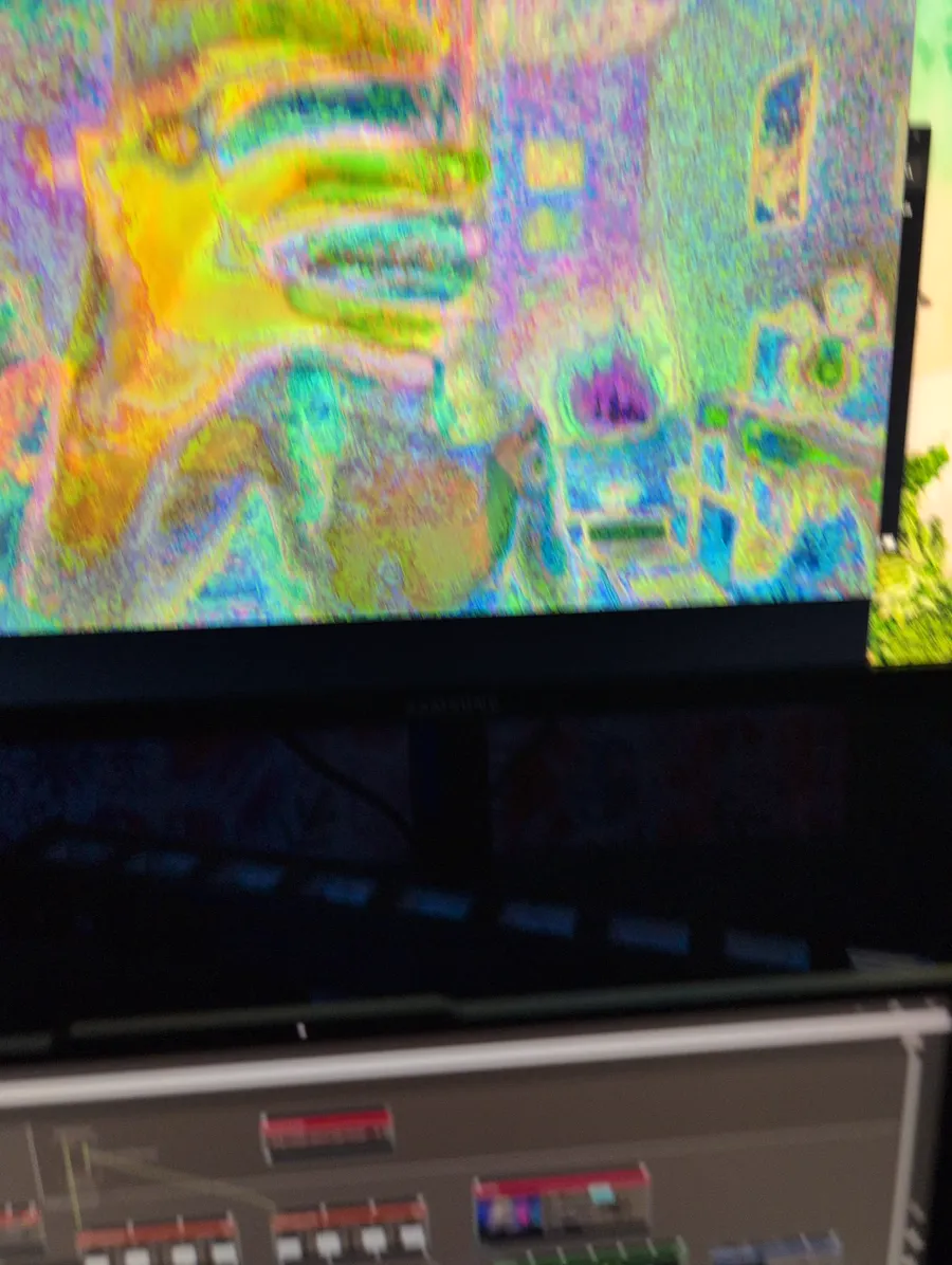 A blurry photo of a large screen full of noisy, rainbow-coloured glitch video, with the Max patch on a laptop below it
