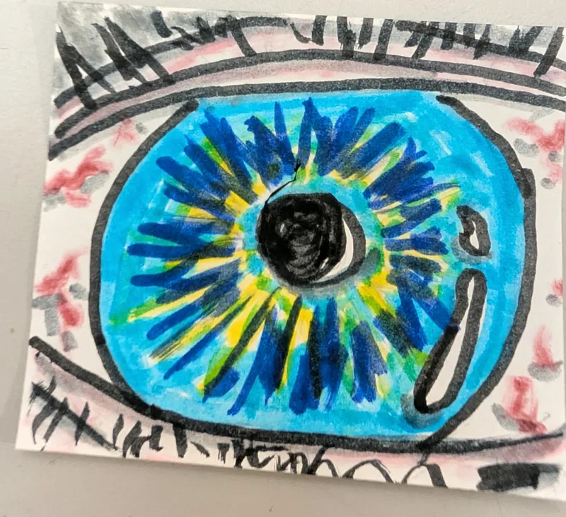 Hand-drawn eye in marker on a post-it note: a bright blue iris with dark blue and yellow rays around a black pupil, lashes above and below