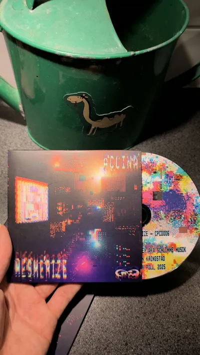 The Mesmerize CD in its sleeve, held in a hand in front of a green watering can