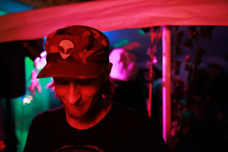 Accina smiling down at the decks under red light, wearing a camouflage cap with an alien patch