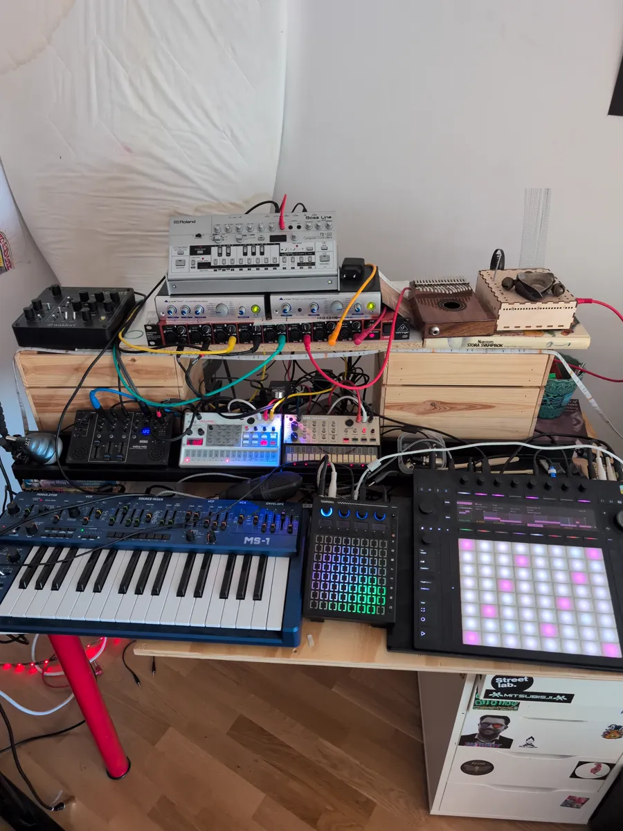 The studio desk: a Roland bass line, Korg Volcas, a Waldorf module, a blue Behringer MS-1 keyboard, a Tempera and an Ableton Push, all patched together with colourful cables
